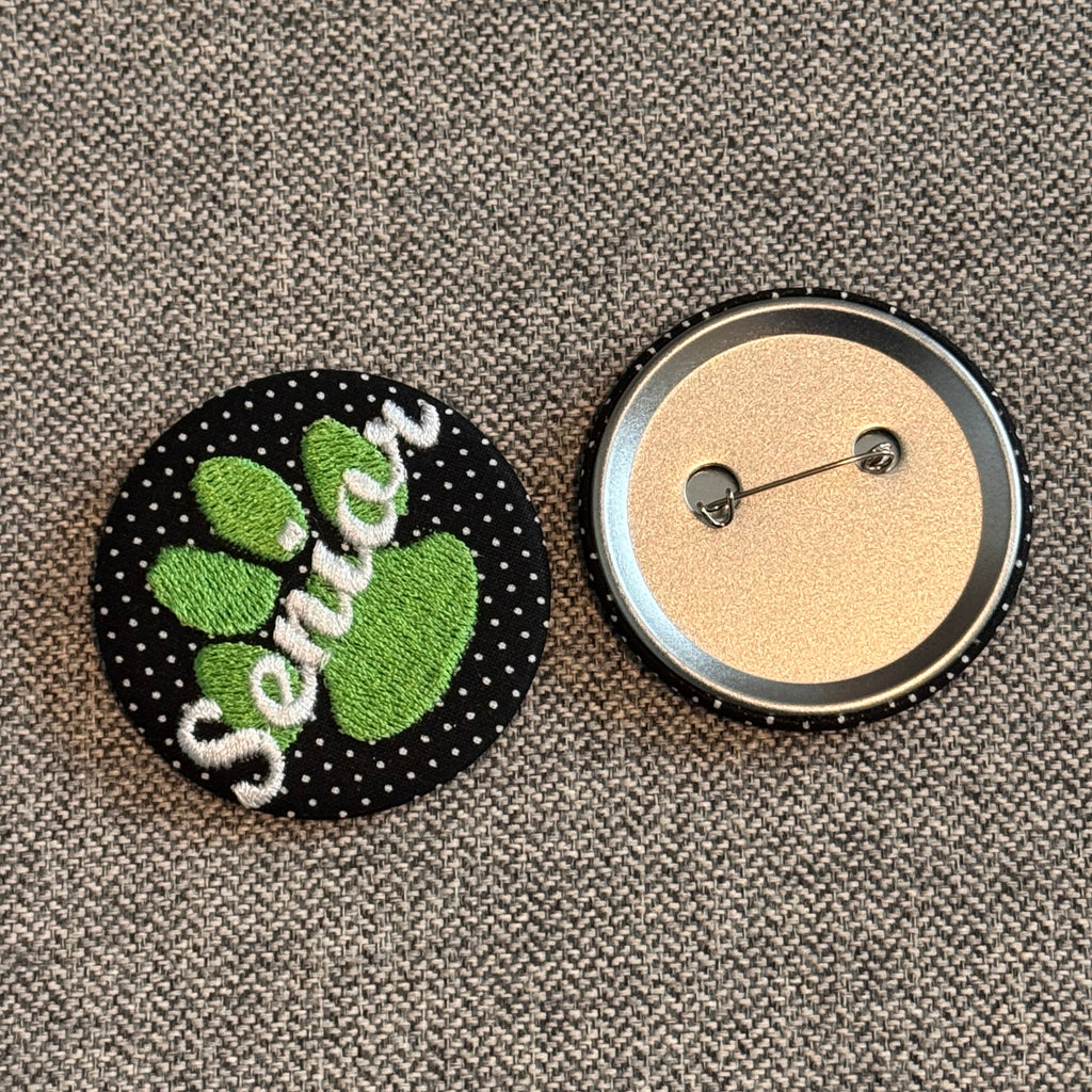Two round buttons on a textured gray fabric background. The top button shows the back with a silver metal rim and pin, and the bottom button shows the front with a black polka-dot background, green paw print, and white embroidered text reading 'Senié'.