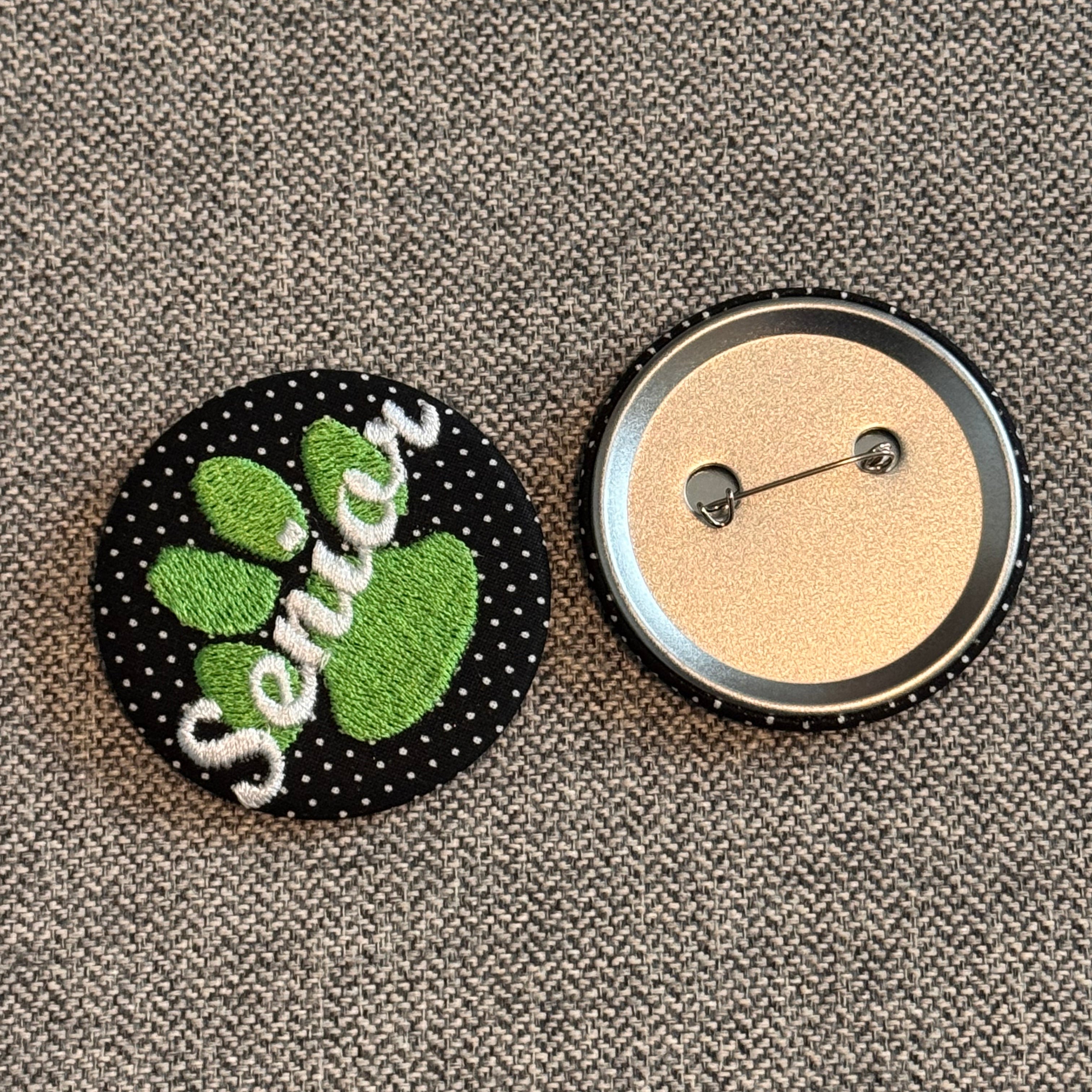 Two round buttons on a textured gray fabric background. The top button shows the back with a silver metal rim and pin, and the bottom button shows the front with a black polka-dot background, green paw print, and white embroidered text reading 'Senié'.