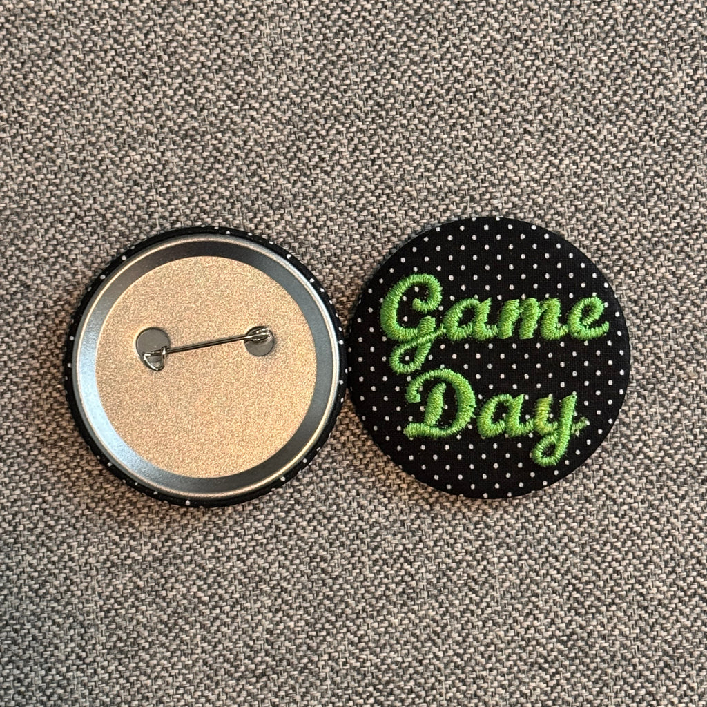Two round pinback buttons on a gray textured fabric background. One button shows the back with a silver metal pin fastener and clear plastic cover over tan glitter fabric.