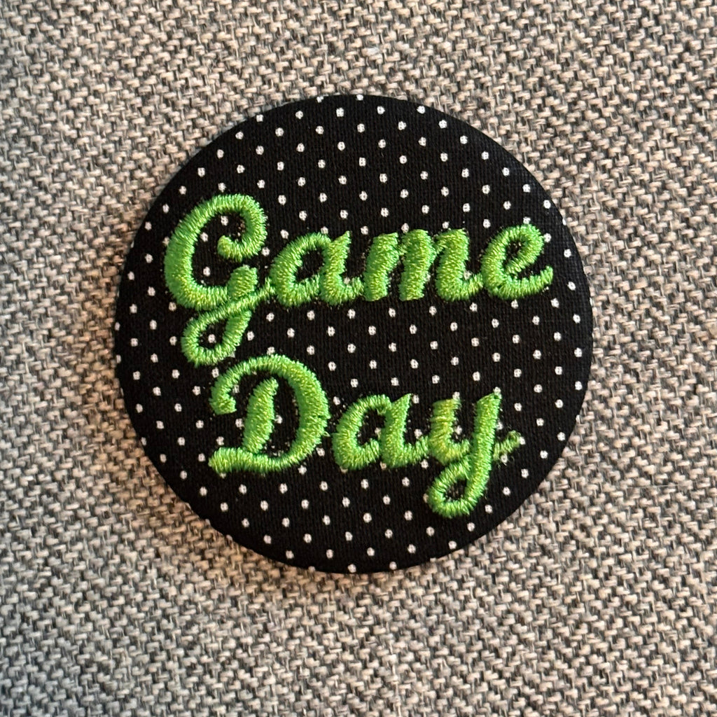 A round black button or patch with white polka dots and the words 'Game Day' embroidered in bright green script, placed on a textured gray fabric background.