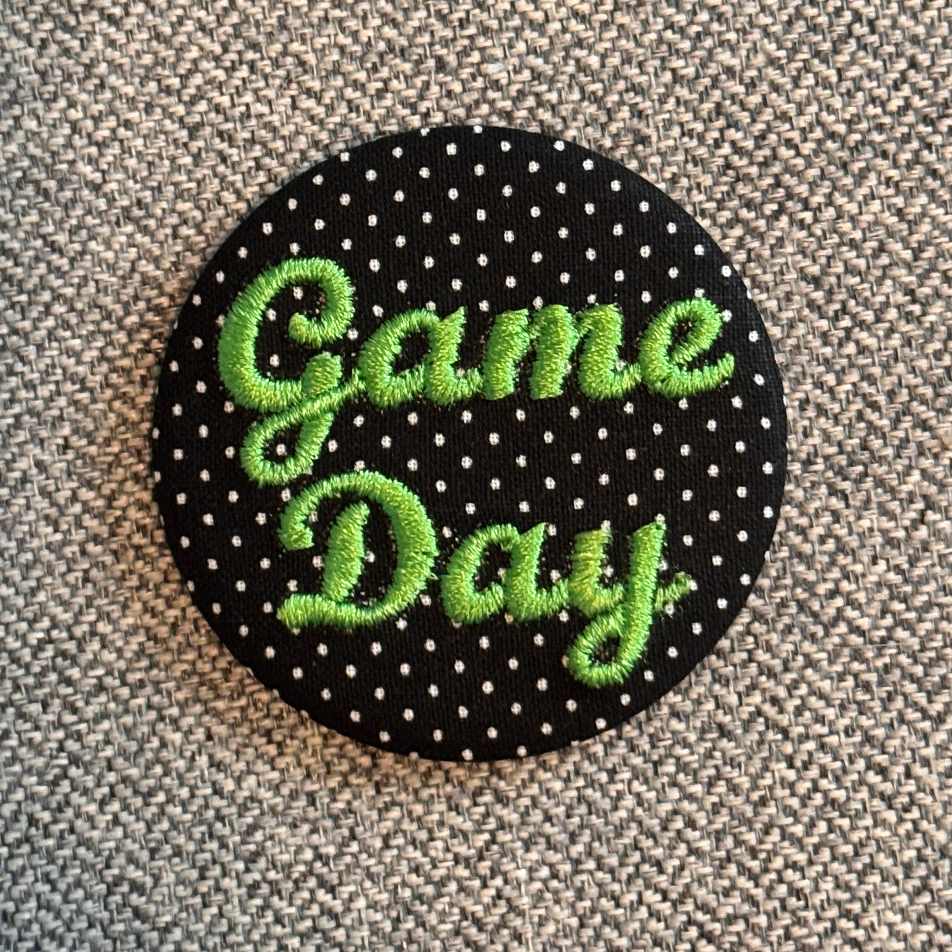 A round black button or patch with white polka dots and the words 'Game Day' embroidered in bright green script, placed on a textured gray fabric background.