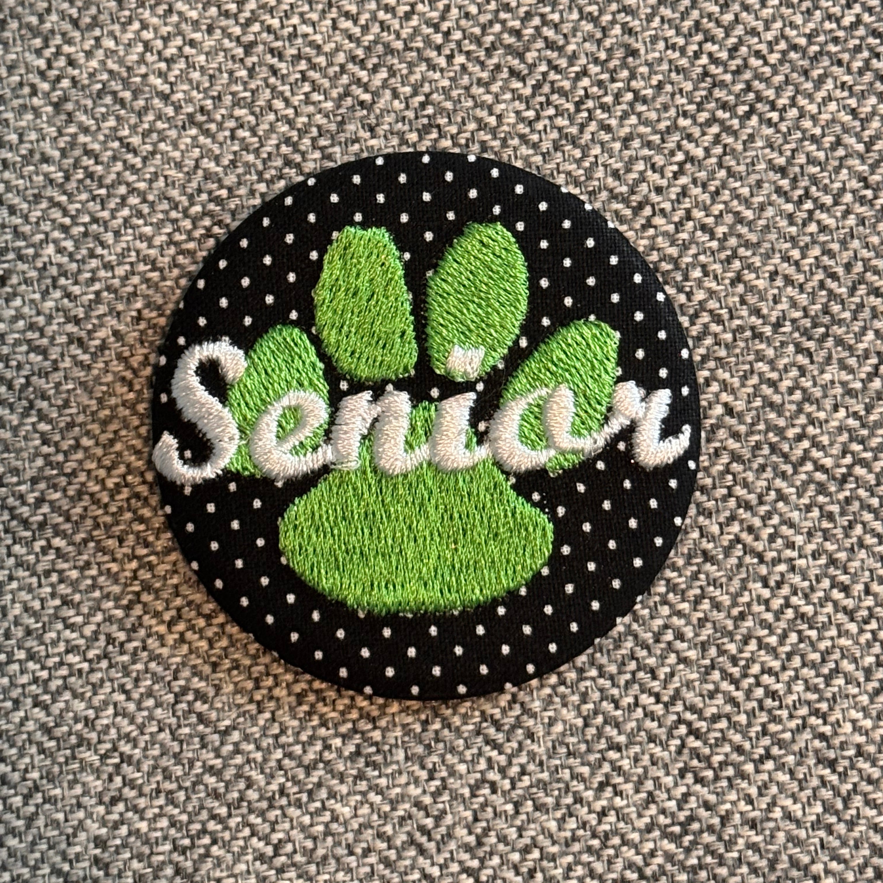 A circular embroidered patch featuring a green paw print over a black background with white polka dots, with the word 'Senior' stitched in white script across the center, placed on a textured gray fabric surface.